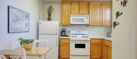 A compact kitchen with wooden cabinets, a white refrigerator, a microwave, and a stove. There is a dining table with chairs and a potted plant.