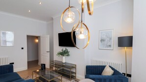 Ein modernes Wohnzimmer mit blauen Sofas, einem Glastisch, einem an der Wand montierten Fernseher und einer Stehlampe.
