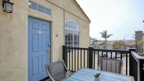 Un balcón con una puerta azul, una mesita con una planta y una silla.