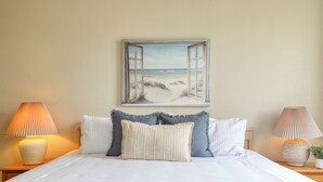 Una cama bien hecha con ropa blanca, dos lámparas de noche y un cuadro con una ventana abierta y vista a la playa.