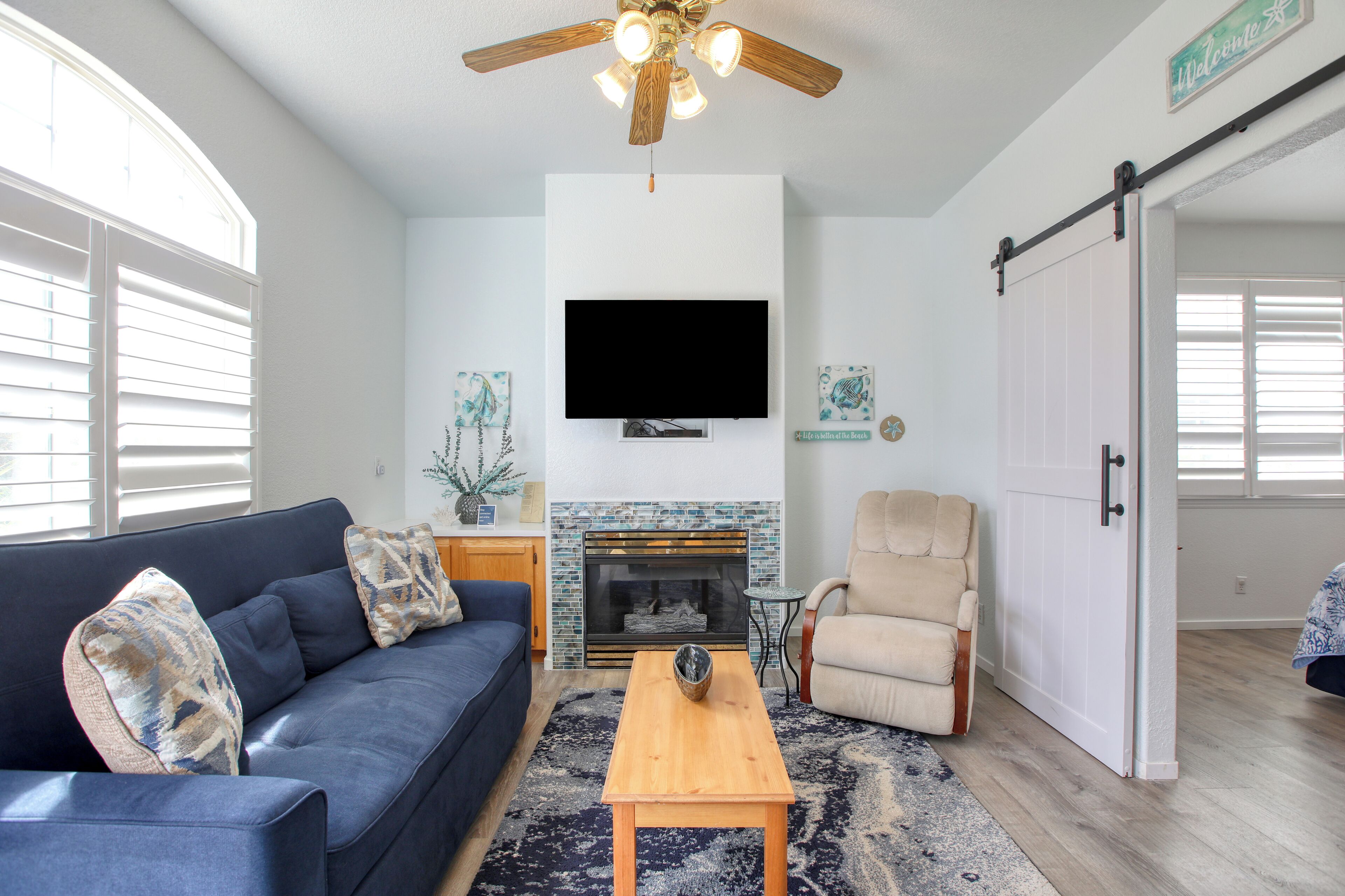 A living room with a blue sofa, a wooden coffee table, a beige armchair, a fireplace, a flat-screen TV, and a ceiling fan.