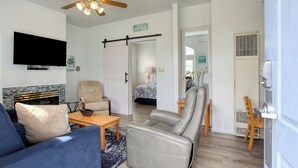 Una sala de estar con sofá, sillón, televisor, ventilador de techo y una puerta que da a un dormitorio.