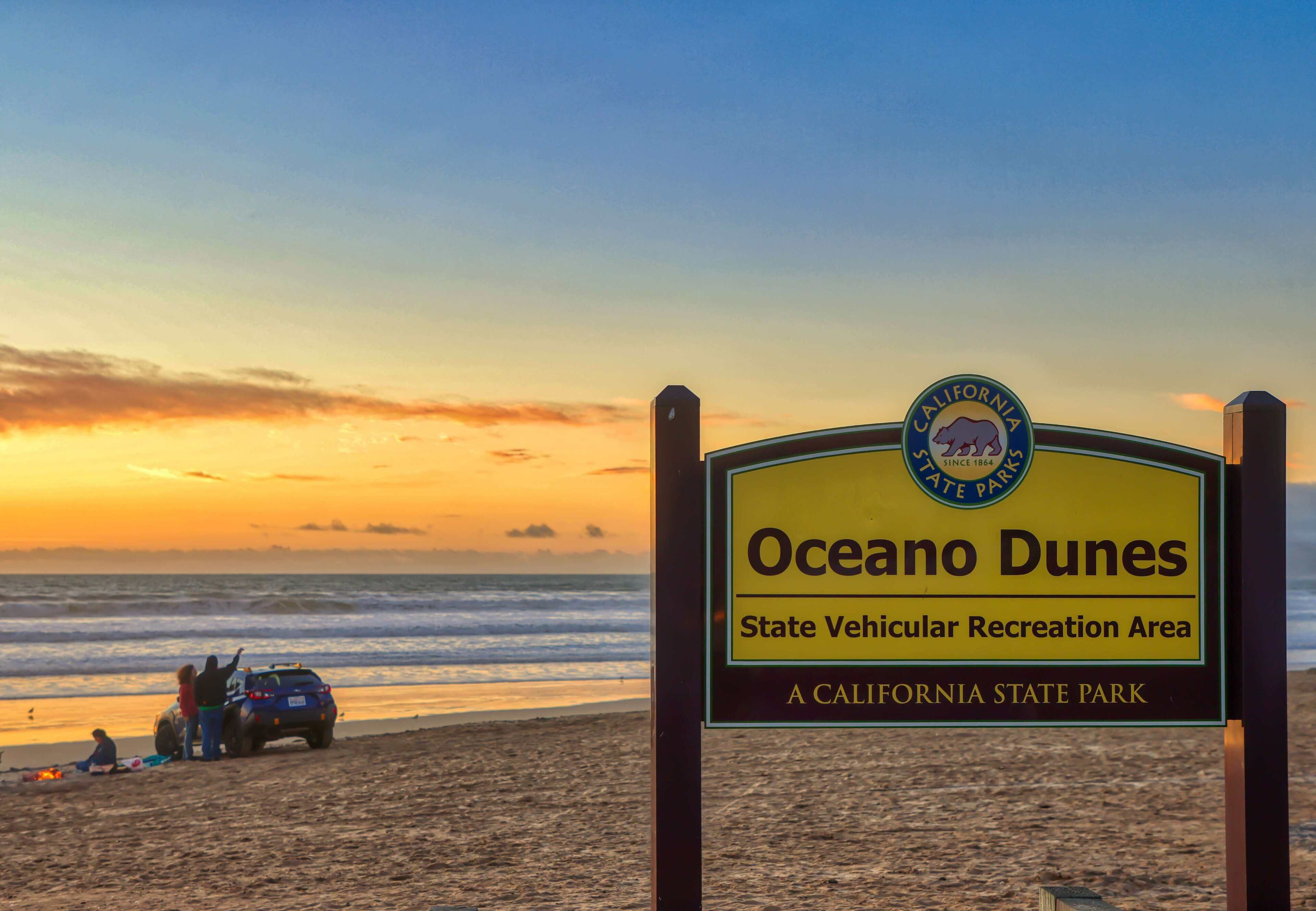 Een strandbordes voor Oceano Dunes State Vehicular Recreation Area, met een zonsondergang op de achtergrond.