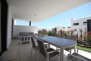 Un patio moderno al aire libre con una mesa de comedor y sillas, un sofá, y vistas a edificios cercanos y un jardín.