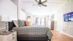 Une chambre à coucher comprenant un lit, une table de chevet, une lampe, un ventilateur de plafond, un téléviseur et une fenêtre avec des rideaux.