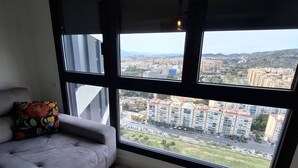 A room with a large window offering a city view, a grey sofa with a colorful cushion, and a wooden floor. - Happy Travel Urban Sky 26A (Málaga)