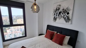 A bedroom with a large bed, a view of buildings through a window, and a wall-mounted abstract art piece. - Happy Travel Urban Sky 26A (Málaga)