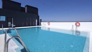 A clear blue pool with metal ladders and red lifebuoys, surrounded by a modern building with a flat roof and a clear sky. - Happy Travel Urban Sky 26A (Málaga)