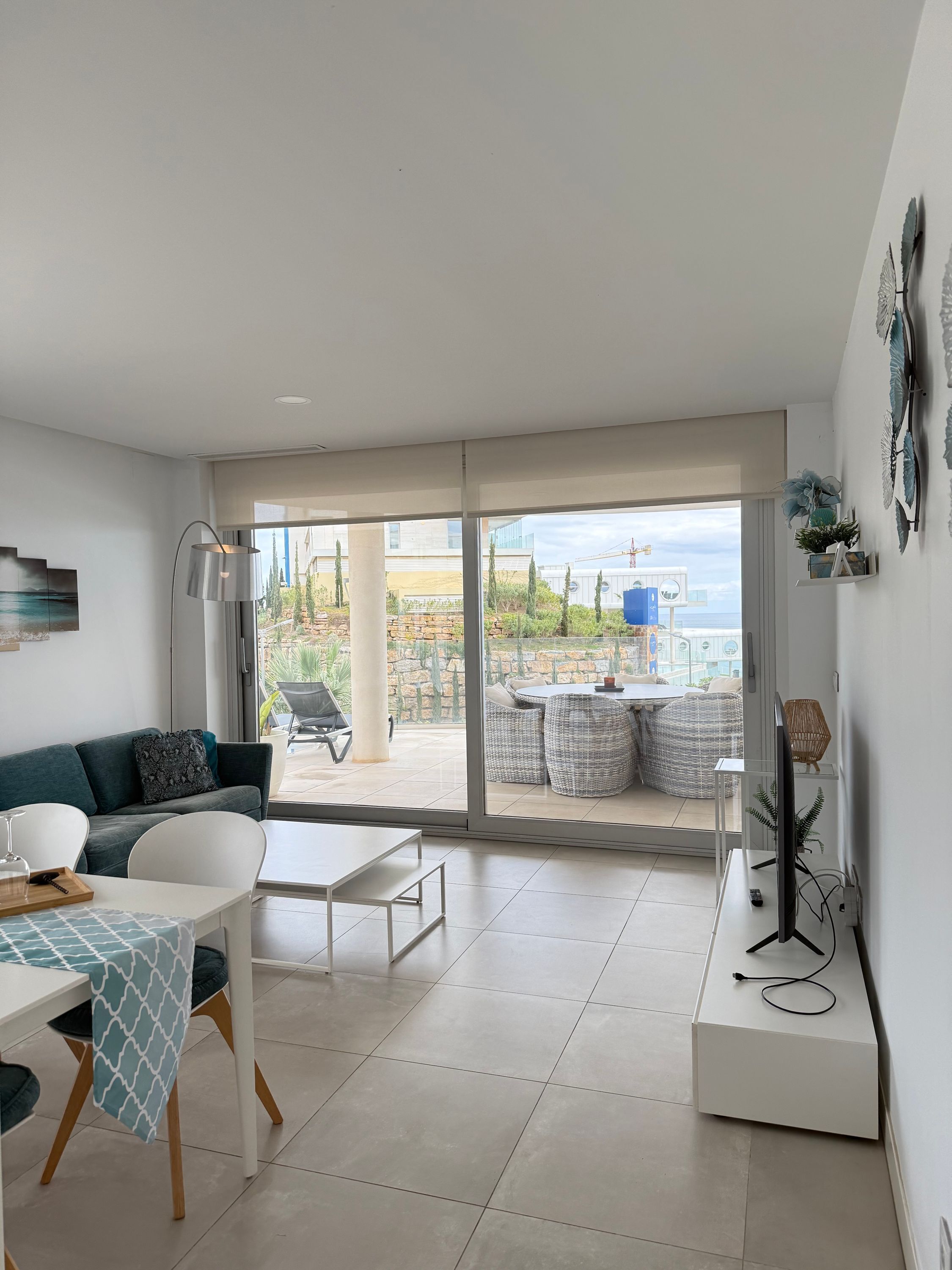 Uma sala de estar moderna com sofá, mesa de jantar e cadeiras. Há uma grande porta de vidro deslizante que dá para um pátio externo com vista para o mar e prédios.