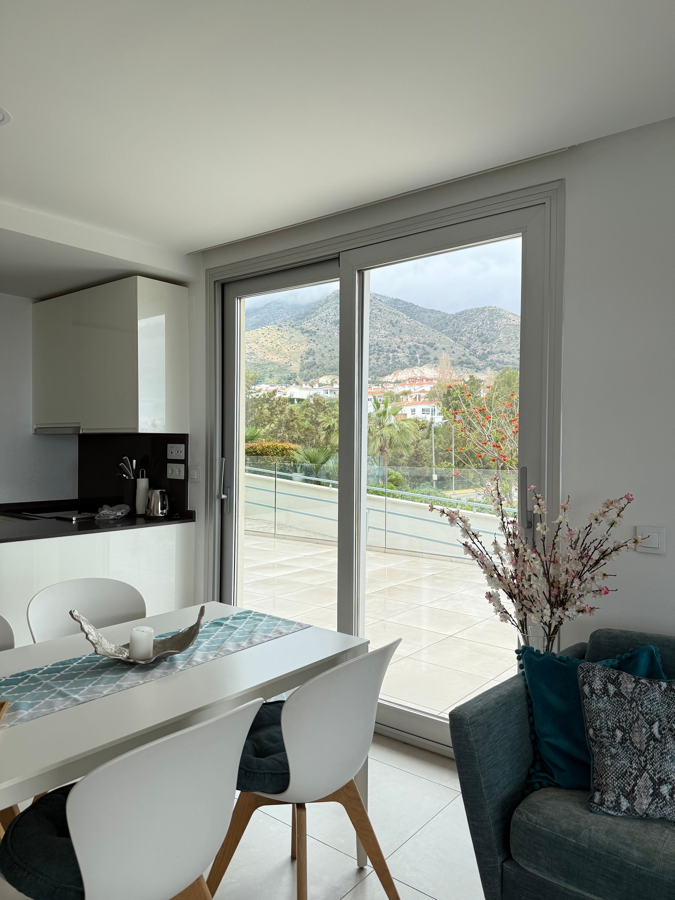 A modern kitchen with white cabinets and a dining area with a table and chairs. A large sliding glass door leads to a balcony with a view of a mountainous landscape.