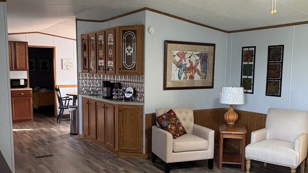 A living room with a wooden floor, a ceiling fan, a coffee table, two armchairs, a side table, and a kitchen area with wooden cabinets and a tiled backsplash.