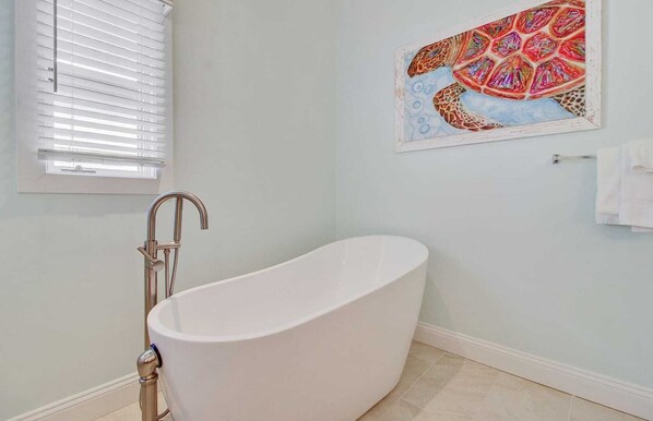 Une salle de bain avec une baignoire sur pieds, une fenêtre avec des stores et une œuvre d’art colorée accrochée au mur.