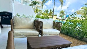 A patio area with a wicker sofa and a wooden table, surrounded by greenery and a building in the background.