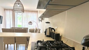 A modern kitchen with a gas stove, a blender, and a kettle. There is a dining area with wicker furniture and a chandelier.