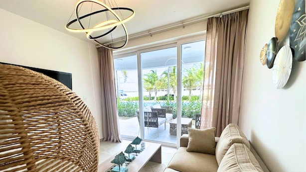 A modern living room with a sofa, a coffee table, and a wicker chair. There is a large window with curtains, and a view of the outdoors.