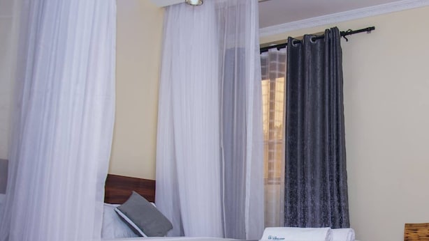A bedroom with a bed, white curtains, a dark curtain, and a hanging light fixture. - The Grand Alton resort (Kisumu)