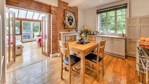 A dining area with a wooden table and chairs, a brick wall, and a large clock. - Spacious rural escape for groups & families (Shropshire)