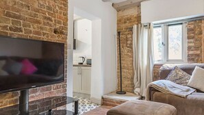 A living room with exposed brick walls, a flat-screen TV, a brown sofa, and a small ottoman. - Spacious rural escape for groups & families (Shropshire)