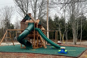 Children can play on the wooden play structure with a slide and swing set amidst a lush, tree-lined area.