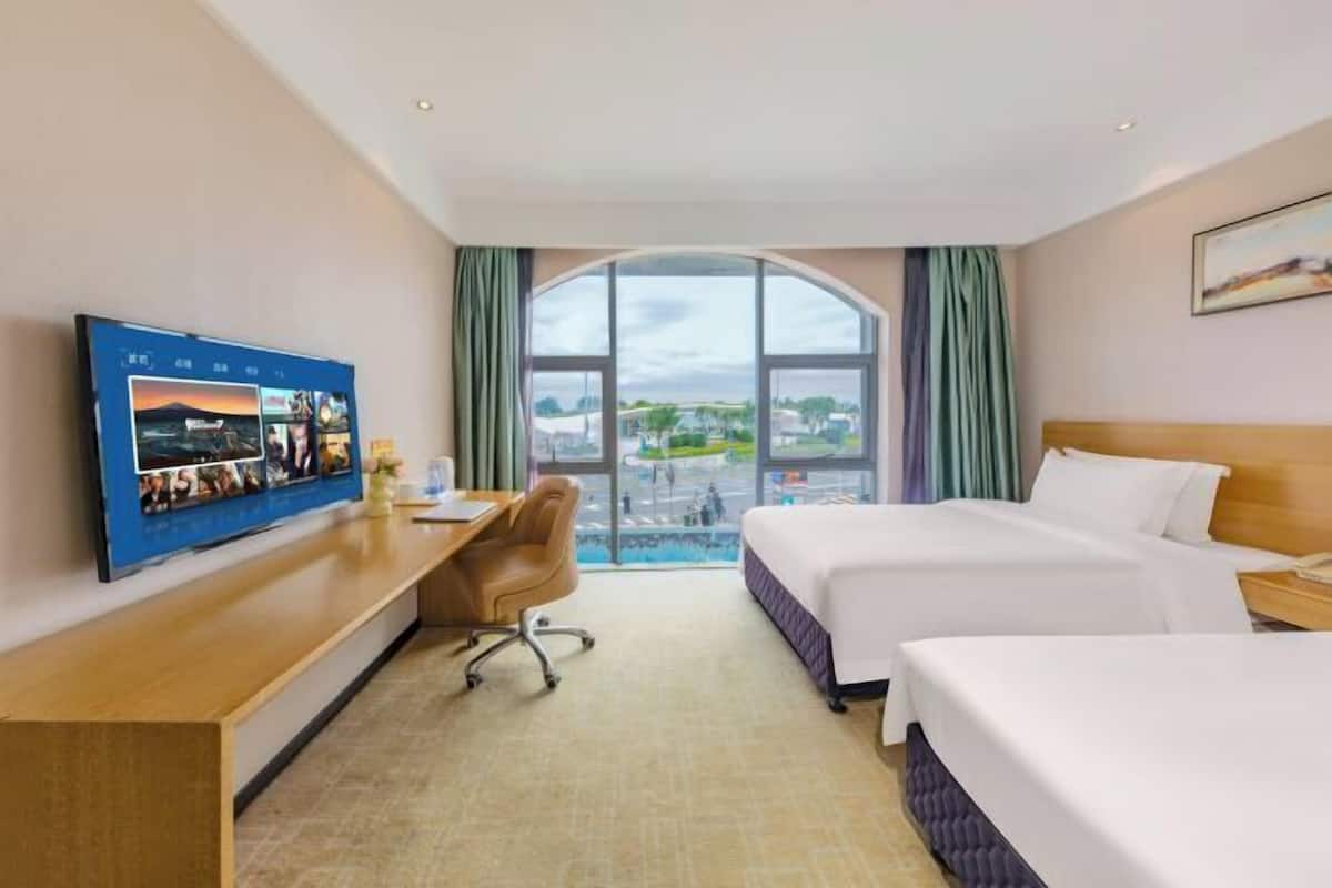 A hotel room with a large window, a desk with a chair, two beds, and a flat-screen TV mounted on the wall.