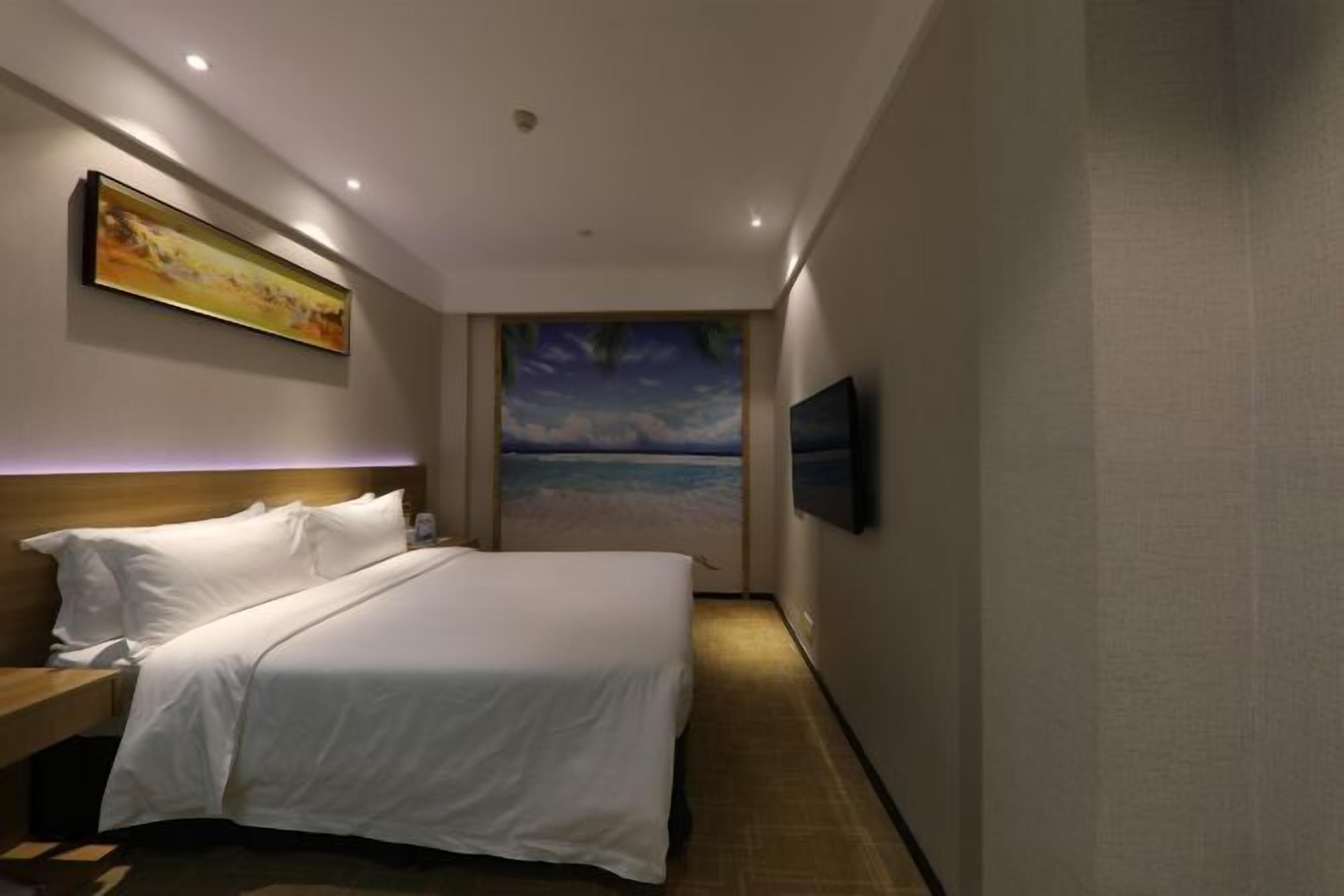 A hotel room with a bed, a painting on the wall, a television, and a view of the beach through a window.
