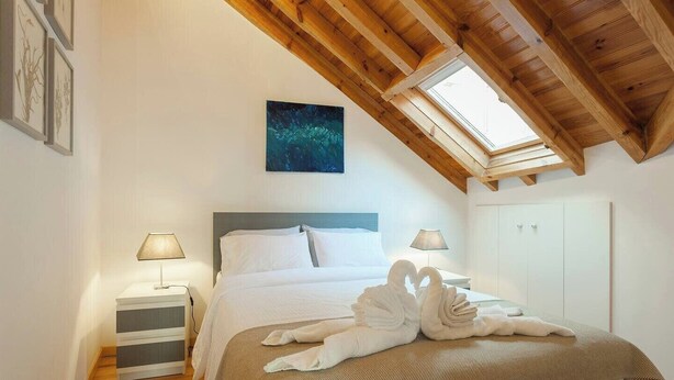 Een slaapkamer met een schuine wand, een bed met witte lakens, twee bedlampen, een schilderij boven het bed en een kledingkast aan de rechterkant.