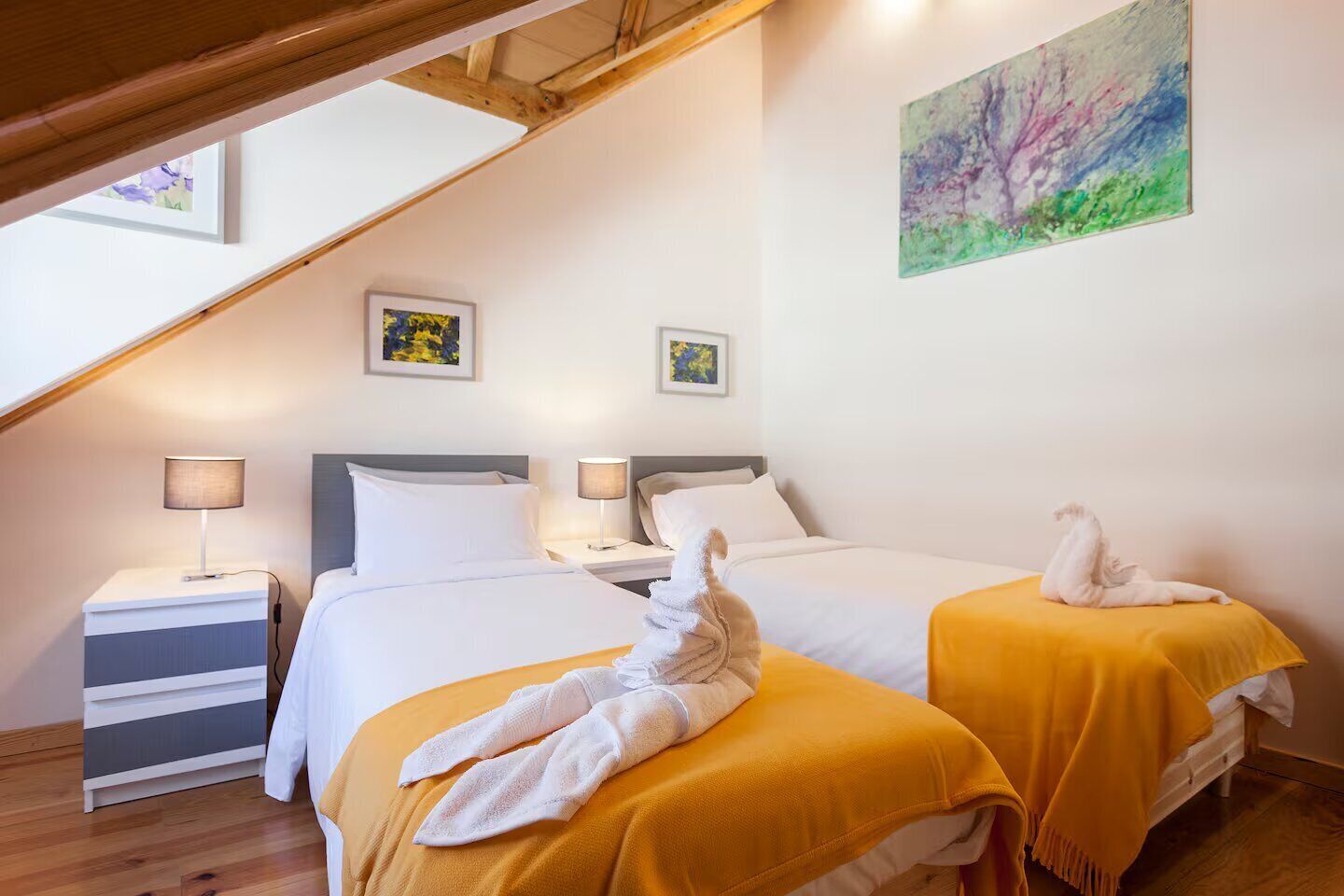 A hotel room with two beds, each with a white duvet and a yellow blanket. There are two bedside tables with lamps, a painting on the wall, and a wooden ceiling beam.