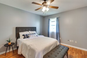 Ein Schlafzimmer mit Bett, Deckenventilator, Fenster mit Vorhängen, einem kleinen Tisch mit Pflanze und einer Bank.