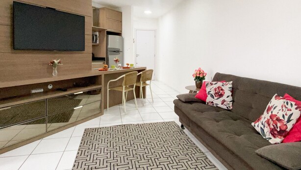 A modern living room with a sofa, a TV mounted on the wall, a dining area with chairs, and a kitchen area with a fridge and cabinets. - Resid. Clube Mar Grosso-Laguna - LAG504 (Laguna)