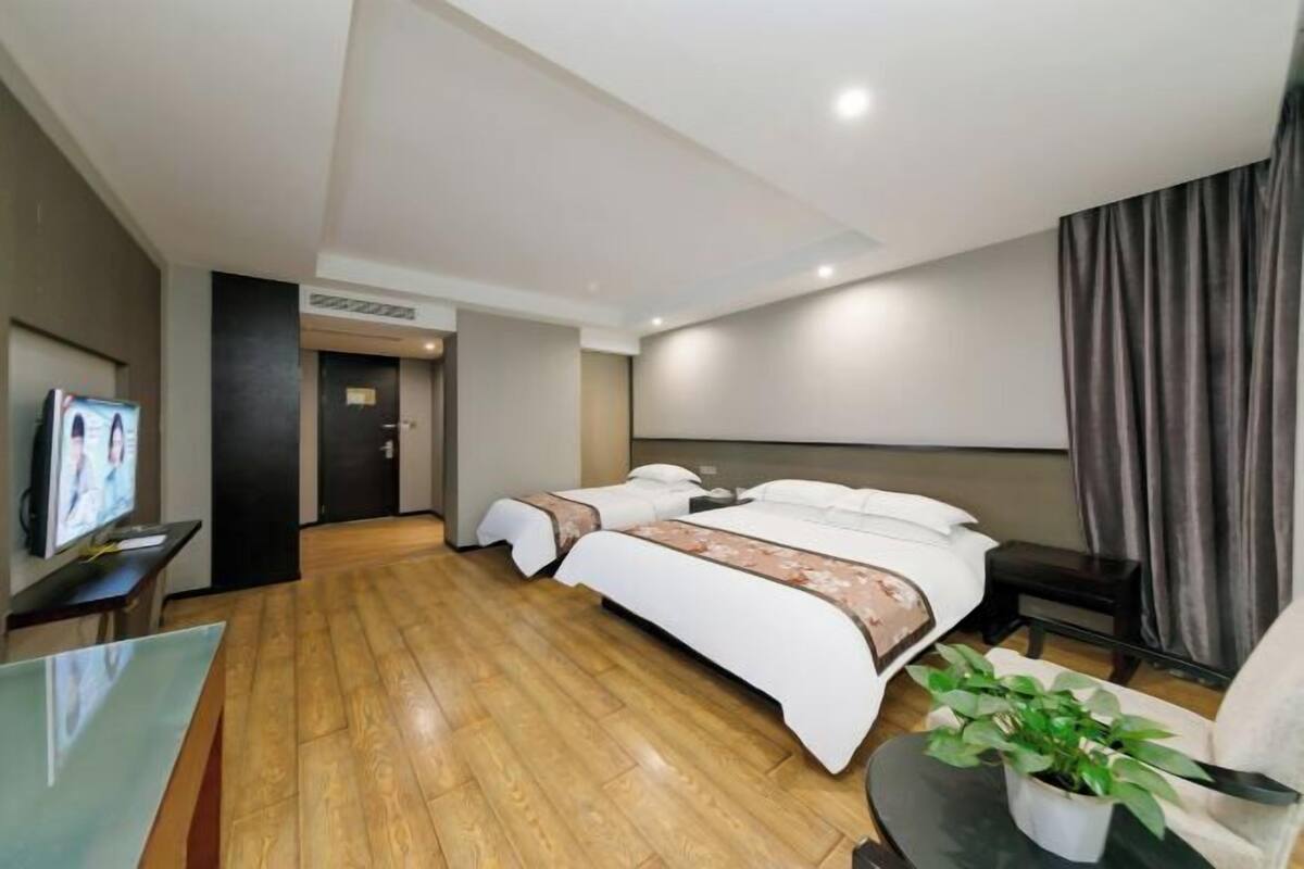 A modern hotel room with a wooden floor, a large bed with white bedding, a TV on a stand, and a small table with a plant.