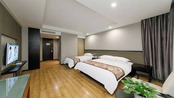 A modern hotel room with a wooden floor, a large bed with white bedding, a TV on a stand, and a small table with a plant.