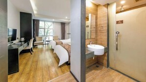 Une chambre d’hôtel moderne avec un plancher en bois, un bureau avec un ordinateur, un lit, une salle de bain avec un lavabo et un miroir, et une grande fenêtre.