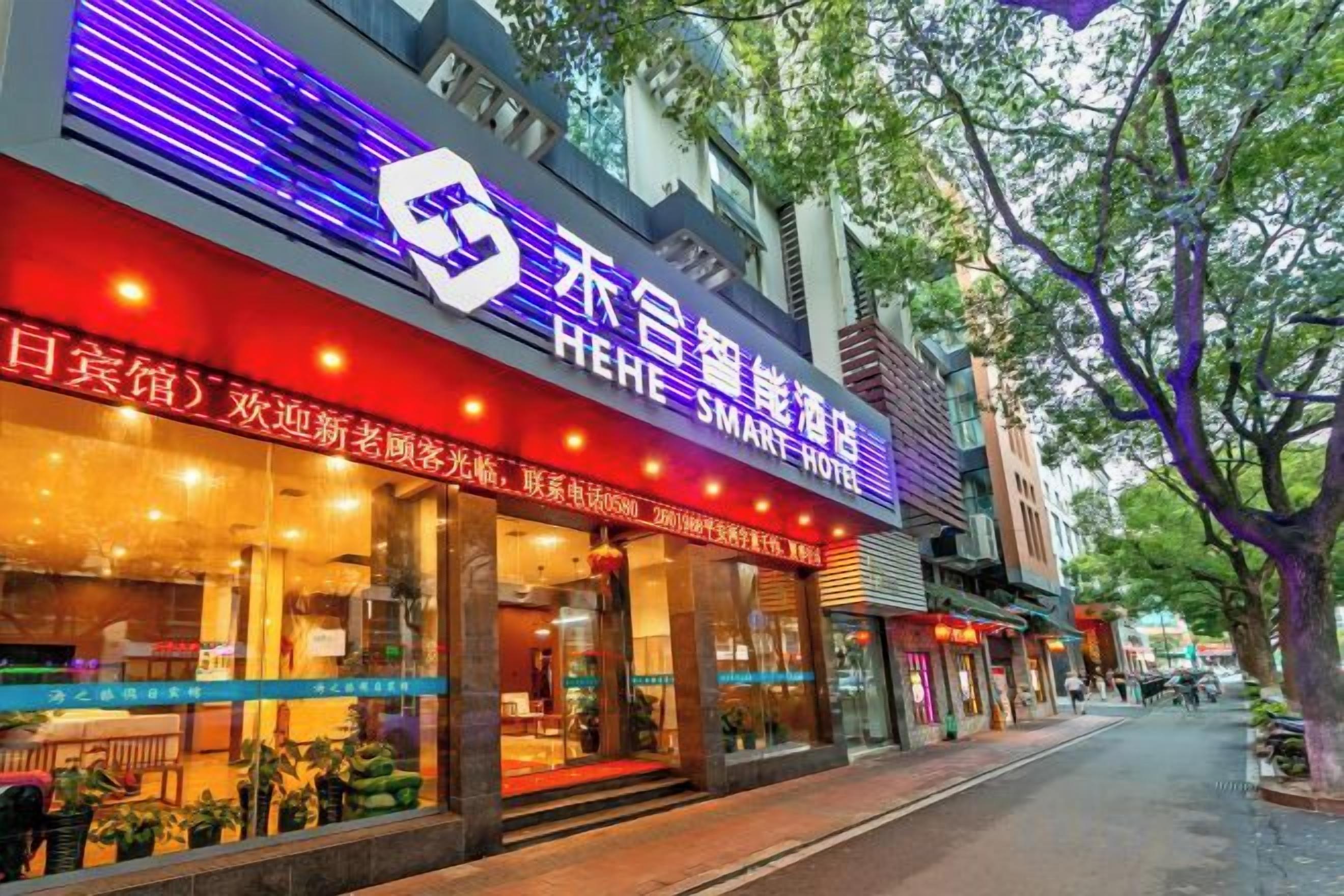 Hotel com placa visível, entrada coberta e vista da rua com árvores e outros edifícios.