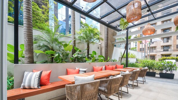 An outdoor dining area with wooden tables and wicker chairs, surrounded by lush greenery and a glass ceiling. - Phoenix suite - Berrini (Cidade Monções São Paulo)
