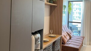 A modern living space with a kitchen area, a dining table with chairs, a sofa, and a large window with a view of buildings. - Flat Makia Beach Experience - Muro Alto (Ipojuca)