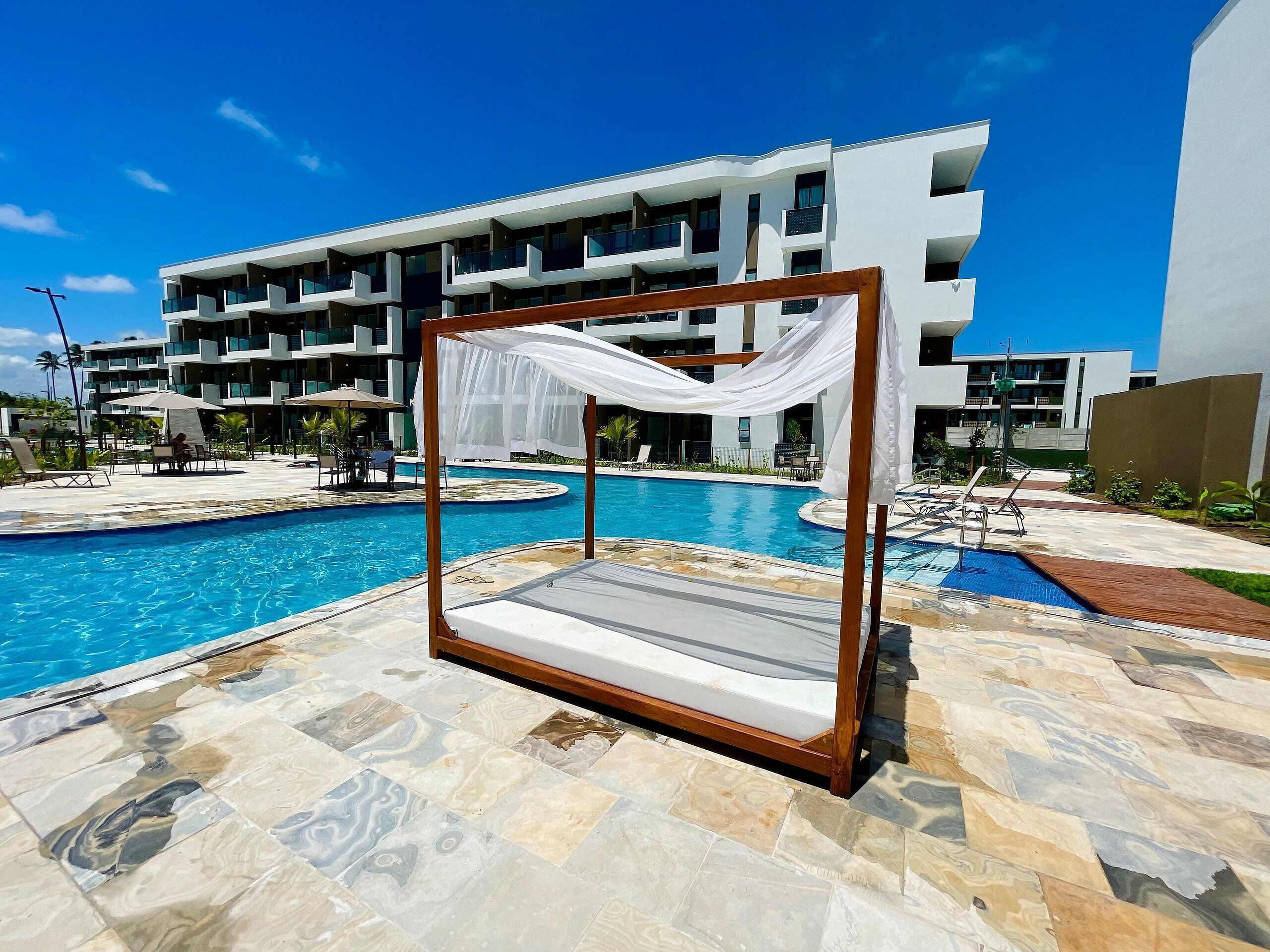 A poolside area with a wooden canopy and a stone-tiled floor, surrounded by a modern building with balconies.