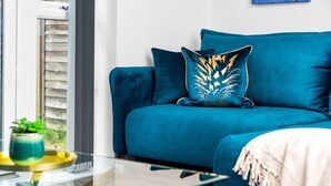 En tealgrøn sofa med en dekorativ pude, et glasbord og et hvidt plaide.