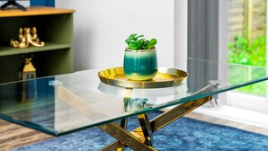 Et moderne interiør med et bord med glasplade, en turkis vase med grønne planter og en guldfarvet bakke.