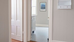 A hallway with wooden flooring and white walls, leading to a kitchen area with a window and a framed picture on the wall.