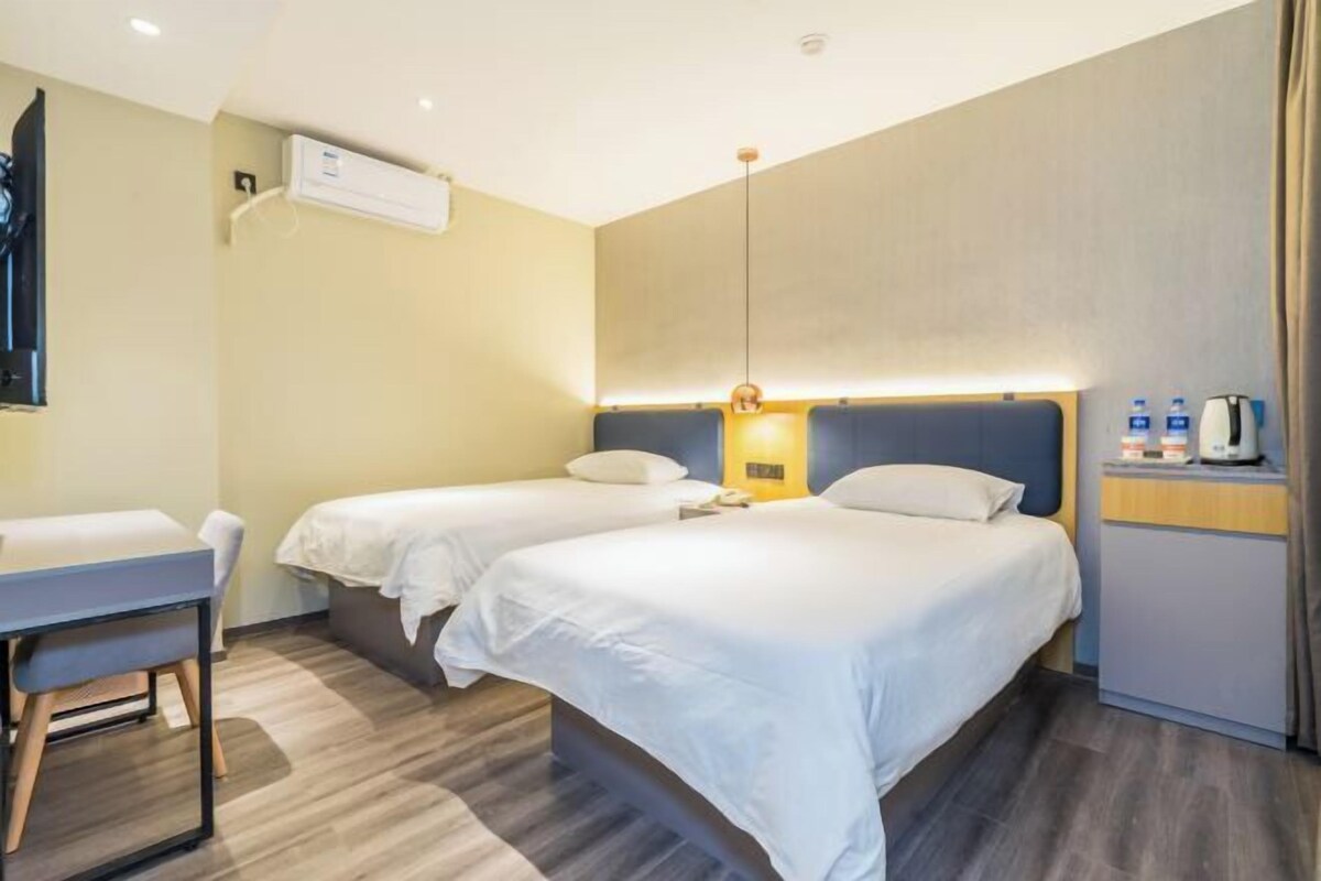 A hotel room with two beds, a desk, and a chair. There is an air conditioning unit on the wall, a hanging light fixture, and a small table with water bottles and a coffee maker.