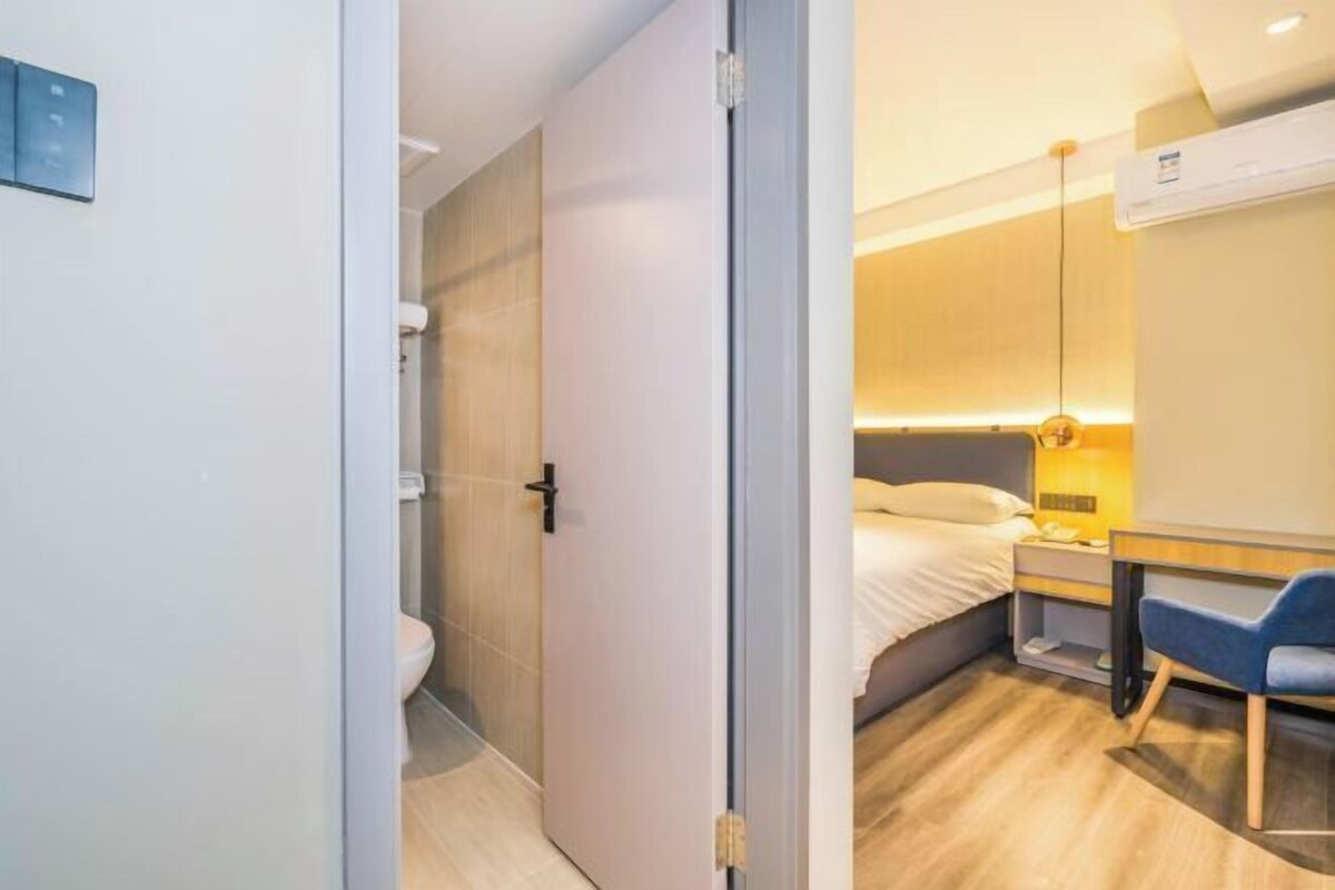 A modern hotel room with a wooden floor, a bed with a headboard, a desk with a chair, and a bathroom with a toilet and shower.