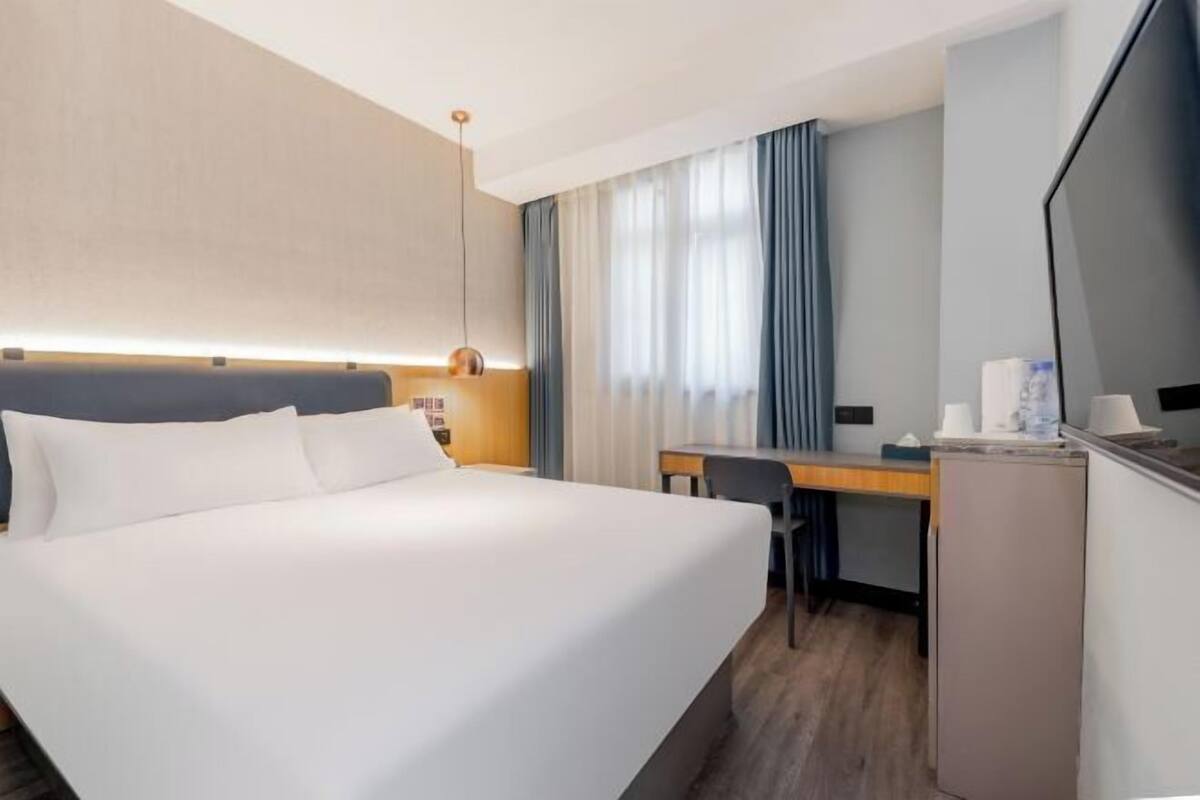 Ett hotellrum med en stor säng, ett skrivbord, en stol, en platt-TV, en minibar och ett fönster med gardiner.