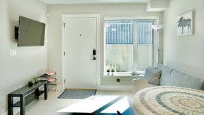 A modern living room with a sofa, a coffee table, a TV mounted on the wall, and a window with a view of the outside.