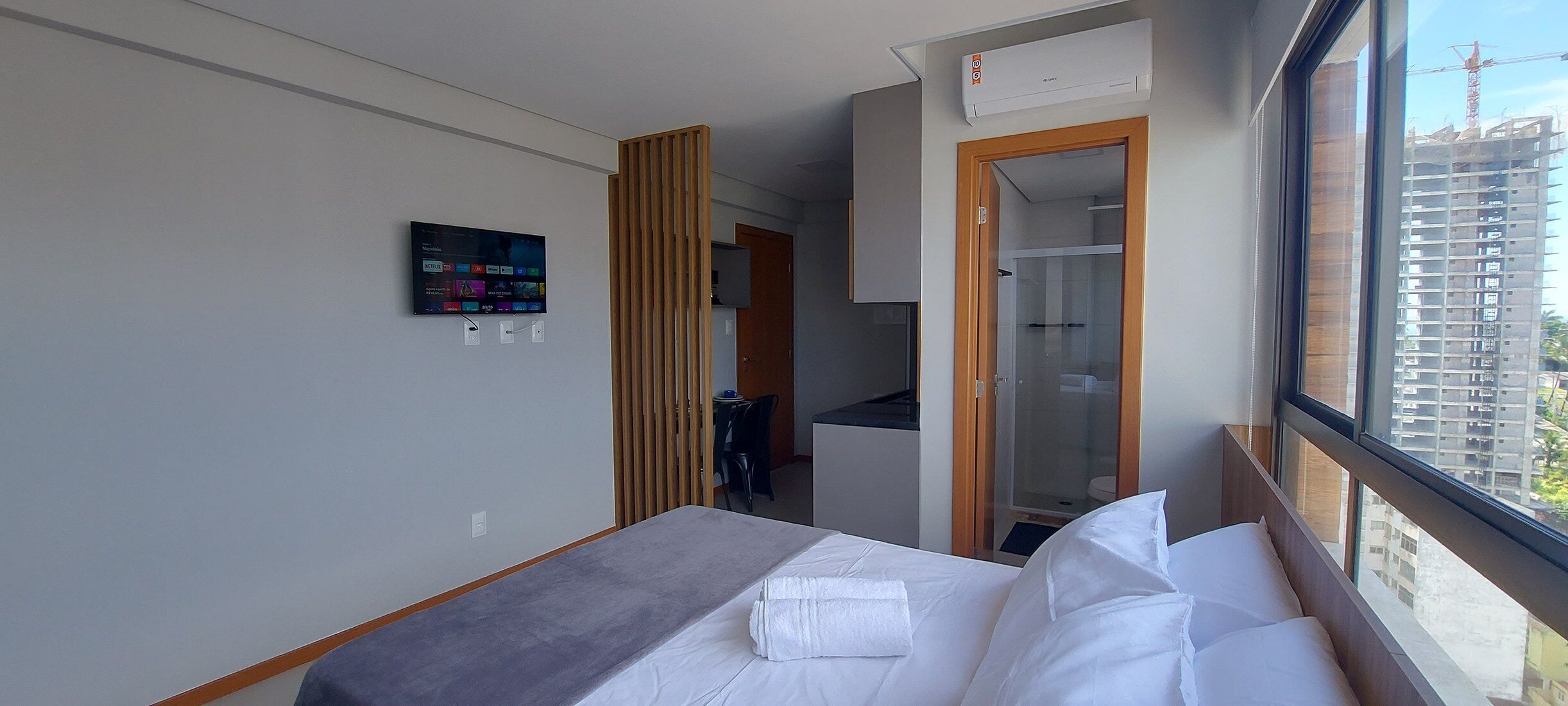 A hotel room with a bed, a TV mounted on the wall, a wooden partition, and a view of a construction site through the window.