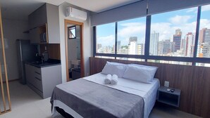 A bedroom with a bed, a nightstand, a desk, and a window with a city view. - COBAR0121 - Edifício Barra Premium (Salvador)
