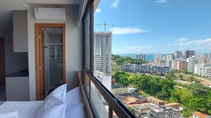 A hotel room with a bed, a desk, and a view of a cityscape with buildings and greenery. - COBAR0121 - Edifício Barra Premium (Salvador)