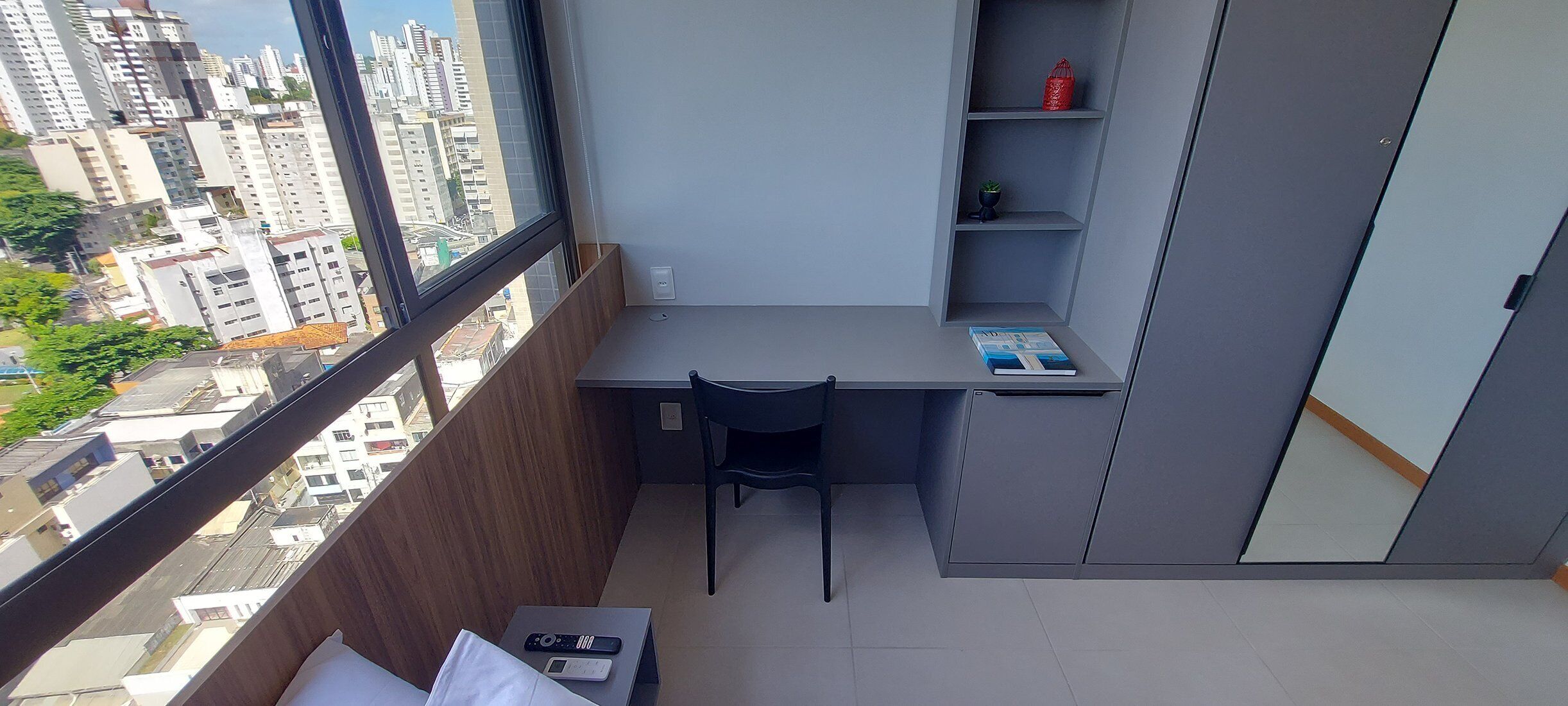 Una habitación pequeña y moderna con escritorio y silla, una estantería y un ventanal con vistas a la ciudad.
