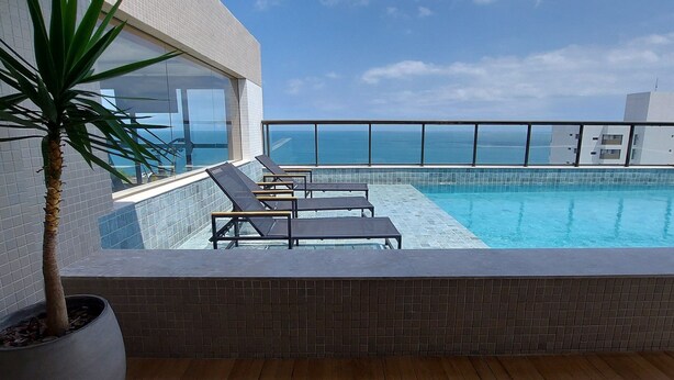 A rooftop pool with lounge chairs, a potted plant, and a view of the ocean. - COBAR0121 - Edifício Barra Premium (Salvador)