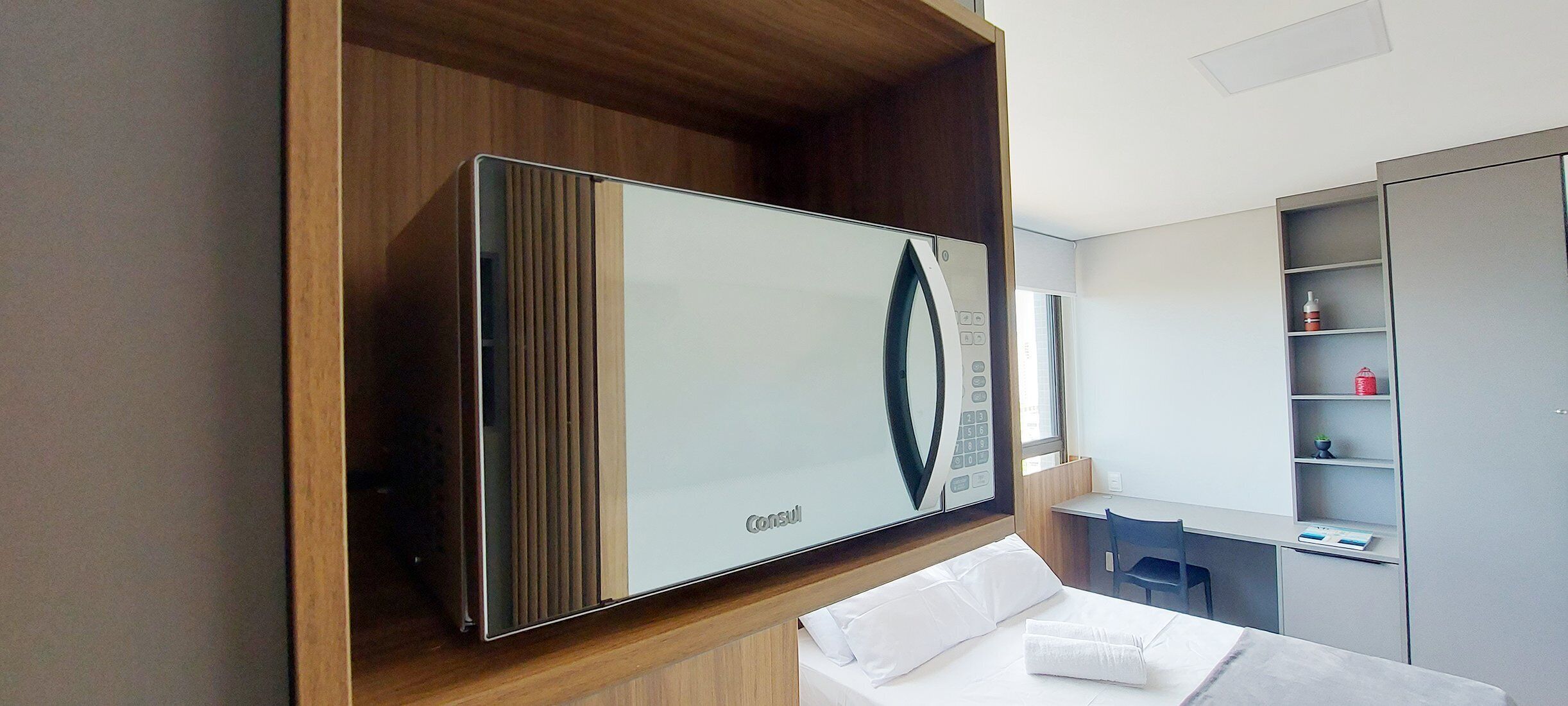 A modern hotel room with a large flat-screen TV mounted on a wooden shelf, a neatly made bed, a small desk with a chair, and a built-in wardrobe with shelves.