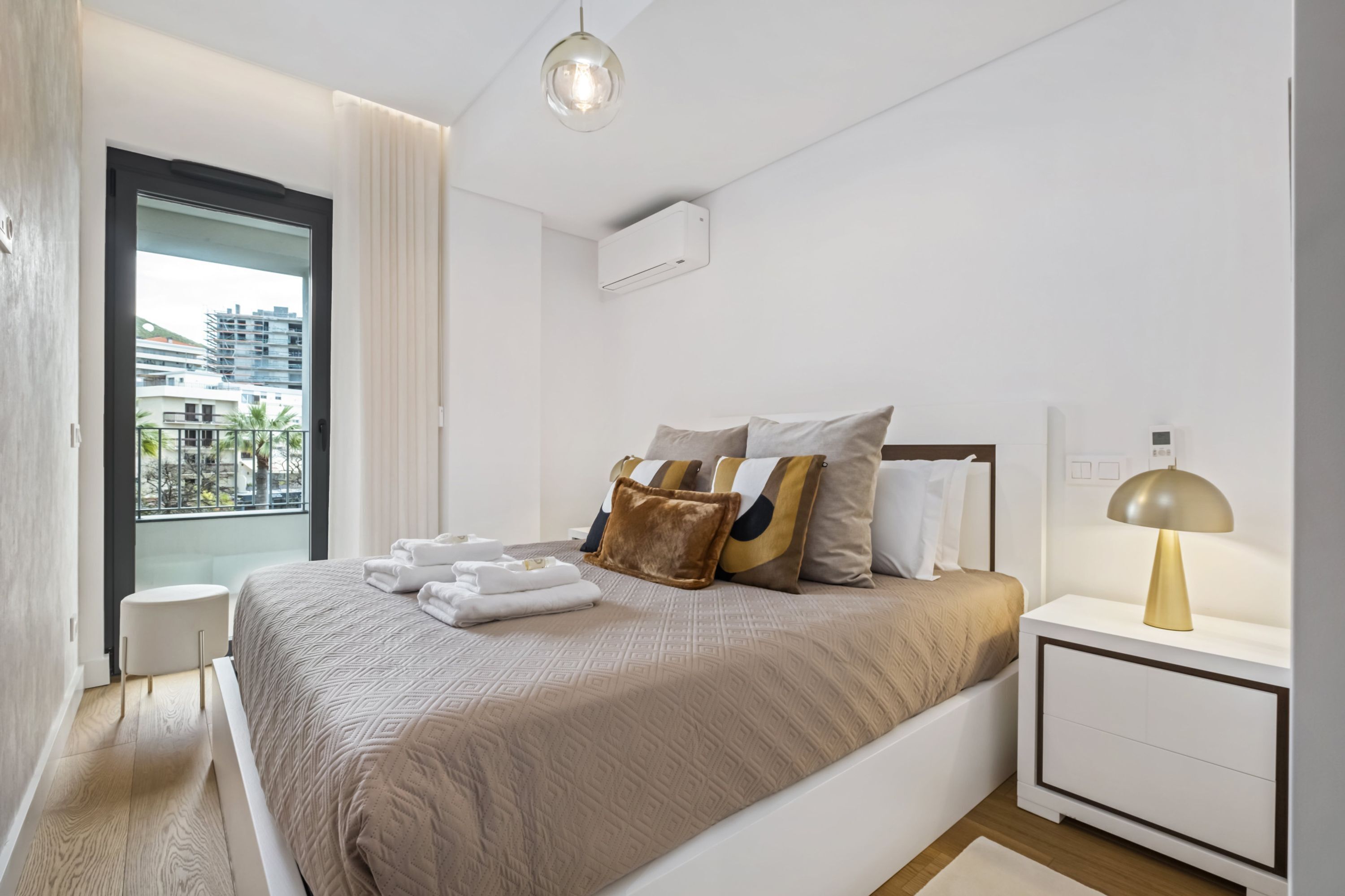 A modern hotel room with a large bed, a bedside table with a lamp, a window with a view of buildings, and a small round table.